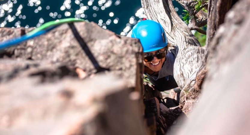 A person wearing safety gear and secured by ropes pauses their climb to look up and smile at the camera. They appear to be high above a body of water. 